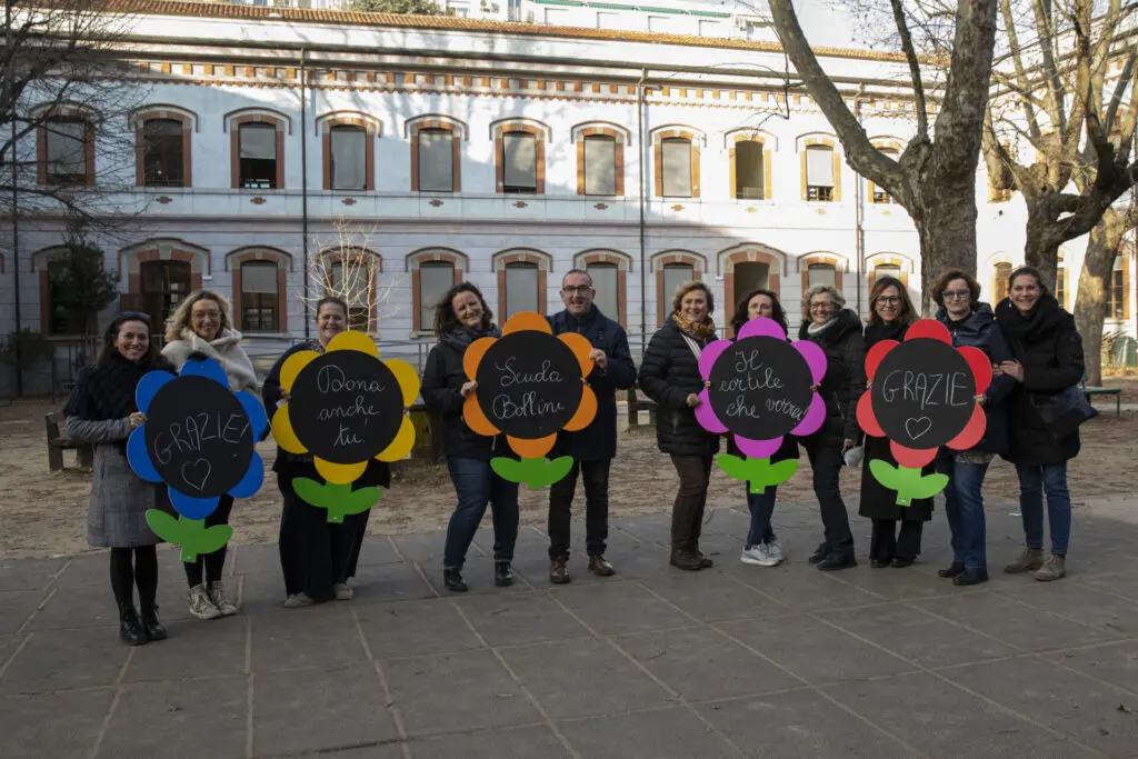 Foto di gruppo di insegnanti in cortile che tengono fiori colorati con messaggi che invitano a donare per il fundraising