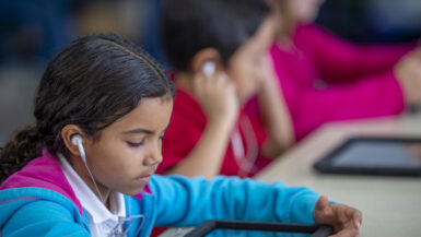 An adorable elementary school girl of Indian ethnicity is using a digital tablet in a classroom with her friends.
