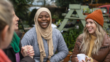Volunteers sitting outdoors wearing warm casual clothing on a sunny cold winters day. They are resting and having a tea break from working on a community farm, looking after crops and performing other sustainable and environmentally friendly tasks. They are laughing and talking together, drinking hot drinks.