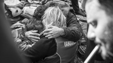 Lesbos, Greece - October 25, 2015: Near the town of Skala Sikamineas on the Greek island of Lesbos, a husband, wife, and their child kneel on the beach overcome with emotion, moments after arriving in a crowded inflatable boat from Turkey. On the right another refugee prepares to light a cigarette.