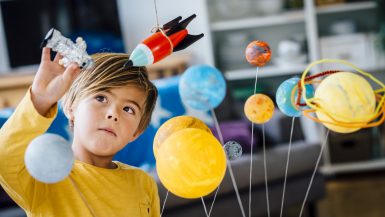 Little boy playing with his homemade planetarium as he holds an astronaut. A rocket hangs above. Arms raised as he plays.