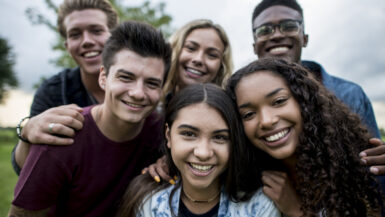 A multi-ethnic group of teenagers are outdoors on a cloudy day. They are wearing casual clothing. They are smiling while taking a selfie together.