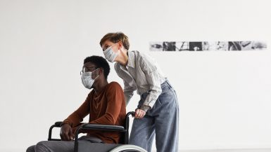 Minimal portrait of African-American man using wheelchair and looking at paintings in modern art gallery with young woman helping him, both wearing masks, copy space
